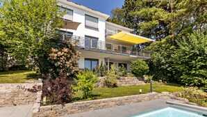 A modern two-story house with a swimming pool, stone retaining walls, and a yellow awning over the balcony. - Apartment 'Am Lichtenberg' with Lake View, Private Terrace and Wi-Fi (Daisendorf)