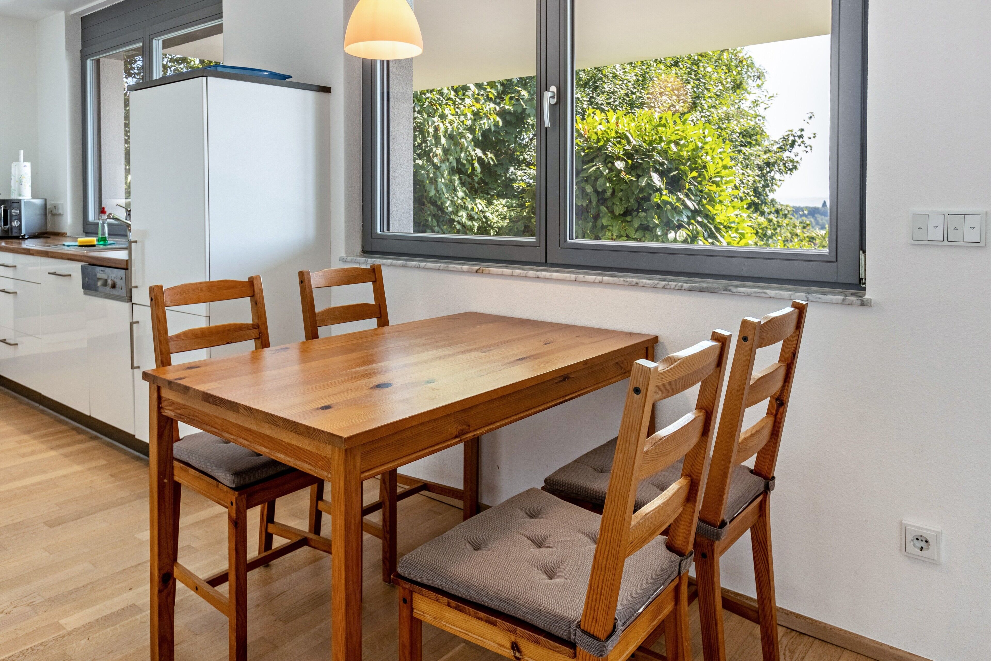 Ein Essbereich mit Holztisch und Stühlen, ein Fenster mit Blick auf Grünflächen und eine Küche im Hintergrund.