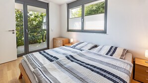 A bedroom with a large bed, a wooden nightstand, a dresser, a window with blinds, and a sliding glass door leading to a balcony. - Apartment 'Am Lichtenberg' with Lake View, Private Terrace and Wi-Fi (Daisendorf)