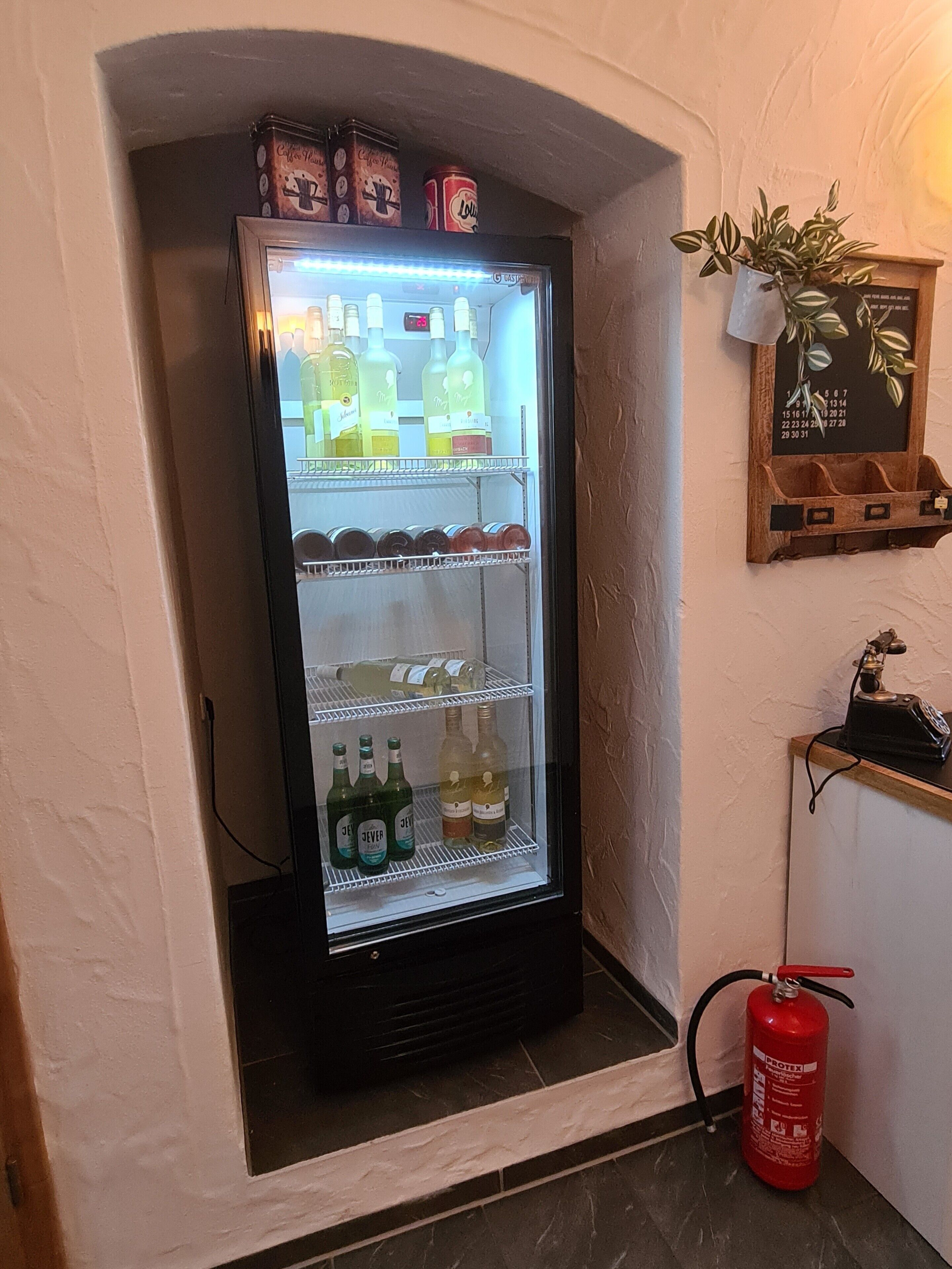 Un refrigerador para bebidas con varias estantes, un extintor y una planta colgada en la pared.
