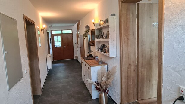 Un pasillo con una puerta de madera, un estante blanco con libros, un florero con plantas secas y un interruptor de luz en la pared.