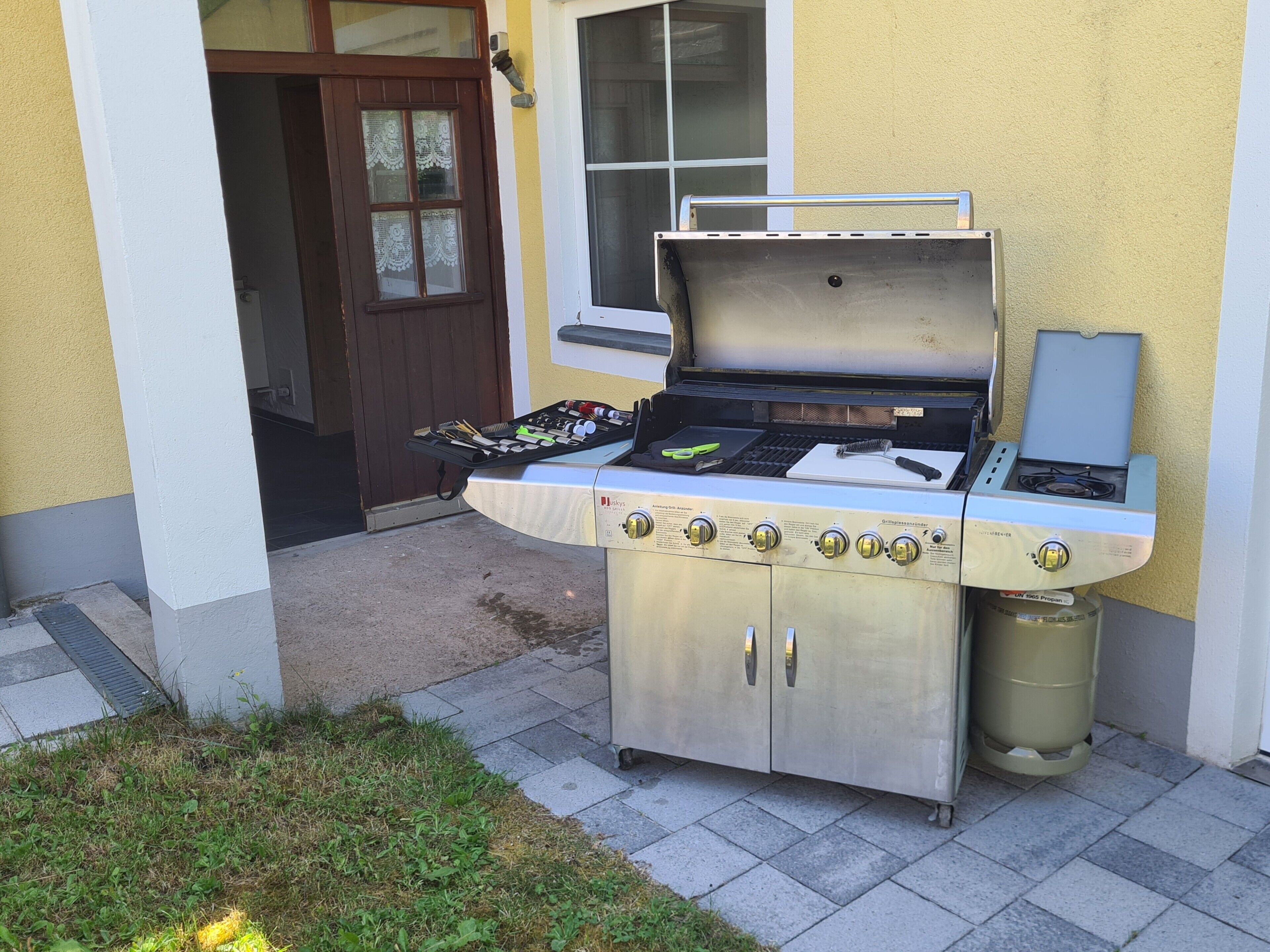 An outdoor gas grill with a side table, a window, and a door in the background.