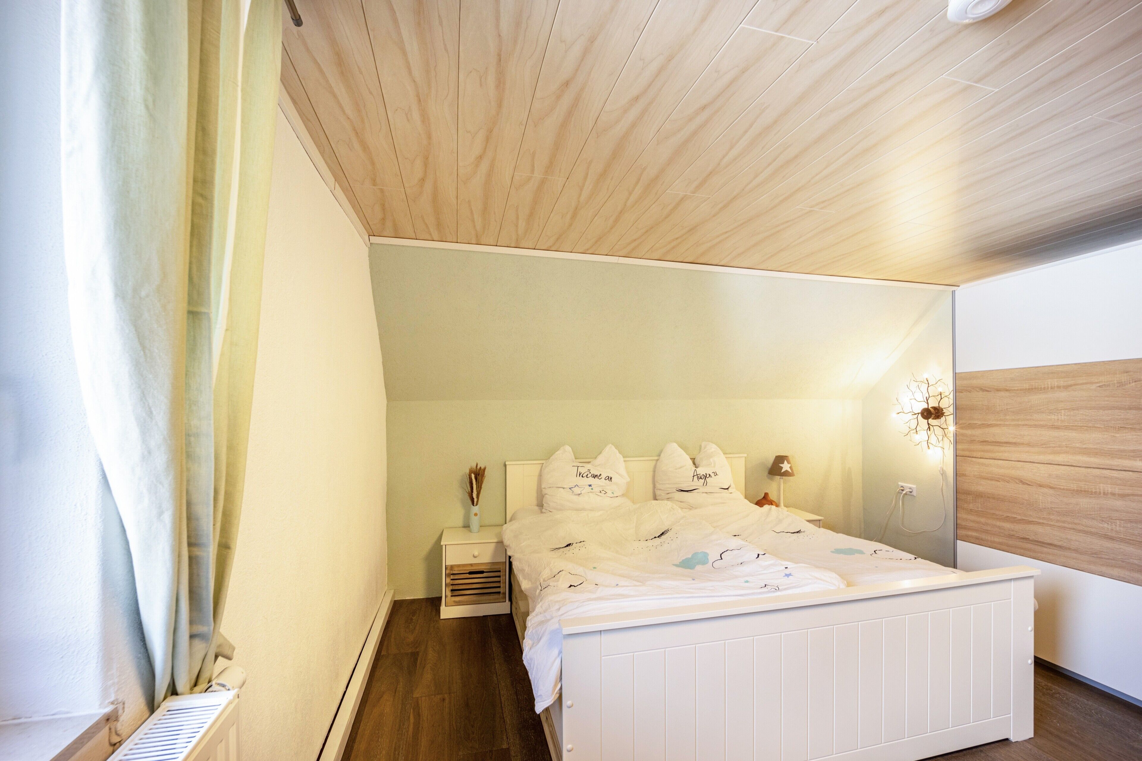 Un dormitorio con techo de madera, una cama blanca con almohadas, una mesita de noche y un aplique de pared.