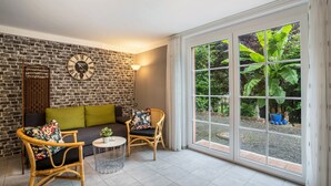 Une pièce avec un mur en briques, un canapé, deux fauteuils en osier, une table ronde et une grande fenêtre donnant sur un paysage verdoyant.