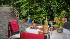 Une table à manger d’extérieur, dressée avec des assiettes, des tasses et une bouteille, entourée de chaises et de verdure.