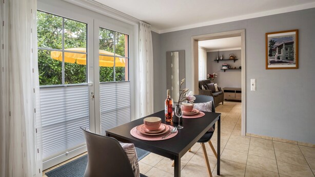 Un coin repas avec une table dressée pour deux personnes, une fenêtre avec des stores et une vue sur la verdure à l’extérieur.