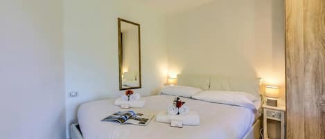 Una cama bien hecha con ropa blanca, una mesita de noche con lámpara y un espejo en la pared.