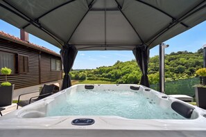 Un jacuzzi con panel de control, rodeado de mobiliario exterior y una zona cubierta, con vistas a un paisaje verde exuberante.