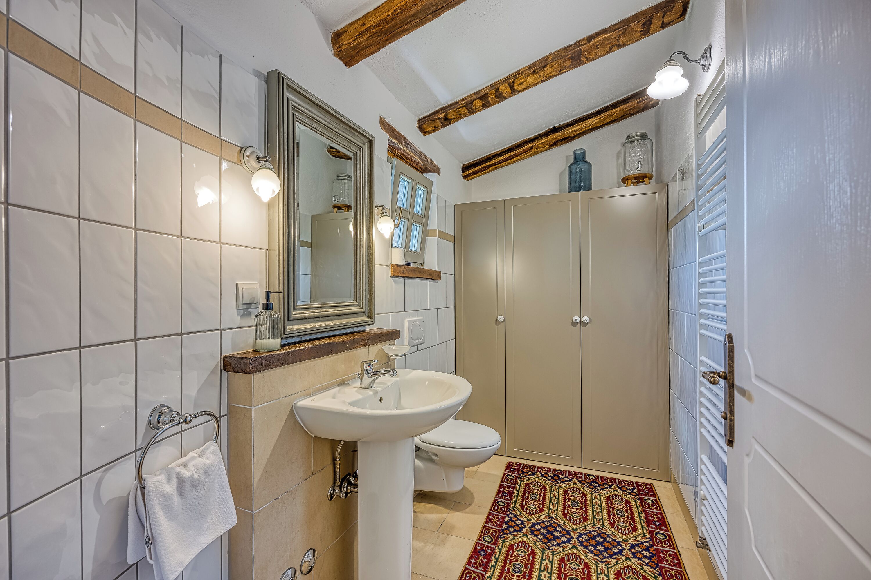 A bathroom with a sink, toilet, and mirror. There is a towel rack and a patterned rug on the floor.