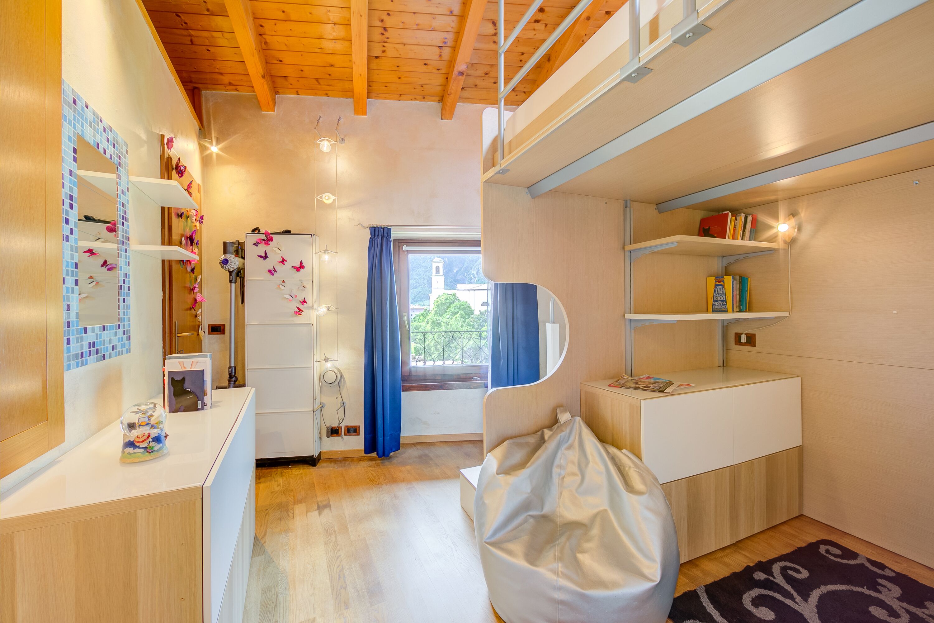 Habitació compacta i ben il·luminada amb un llit superposat, un escriptori amb un ordinador i una finestra amb cortines.