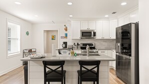 A modern kitchen with white cabinets, stainless steel appliances, and a central island with two bar stools. - Sunflower Hideaway -Walk to eats, bike to beach, pet friendly, Relaxed backyard. (St. Simons Island)