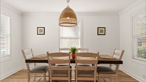 A dining area with a wooden table, four chairs, a woven pendant light, and two framed pictures on the walls. - Sunflower Hideaway -Walk to eats, bike to beach, pet friendly, Relaxed backyard. (St. Simons Island)