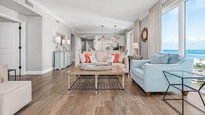 A spacious living area with a light blue sofa, a wooden coffee table, and a view of the city through large windows. - Beautiful 5 Star Condo right on the Beach!  Breathtaking Views!! (Navarre Beach)