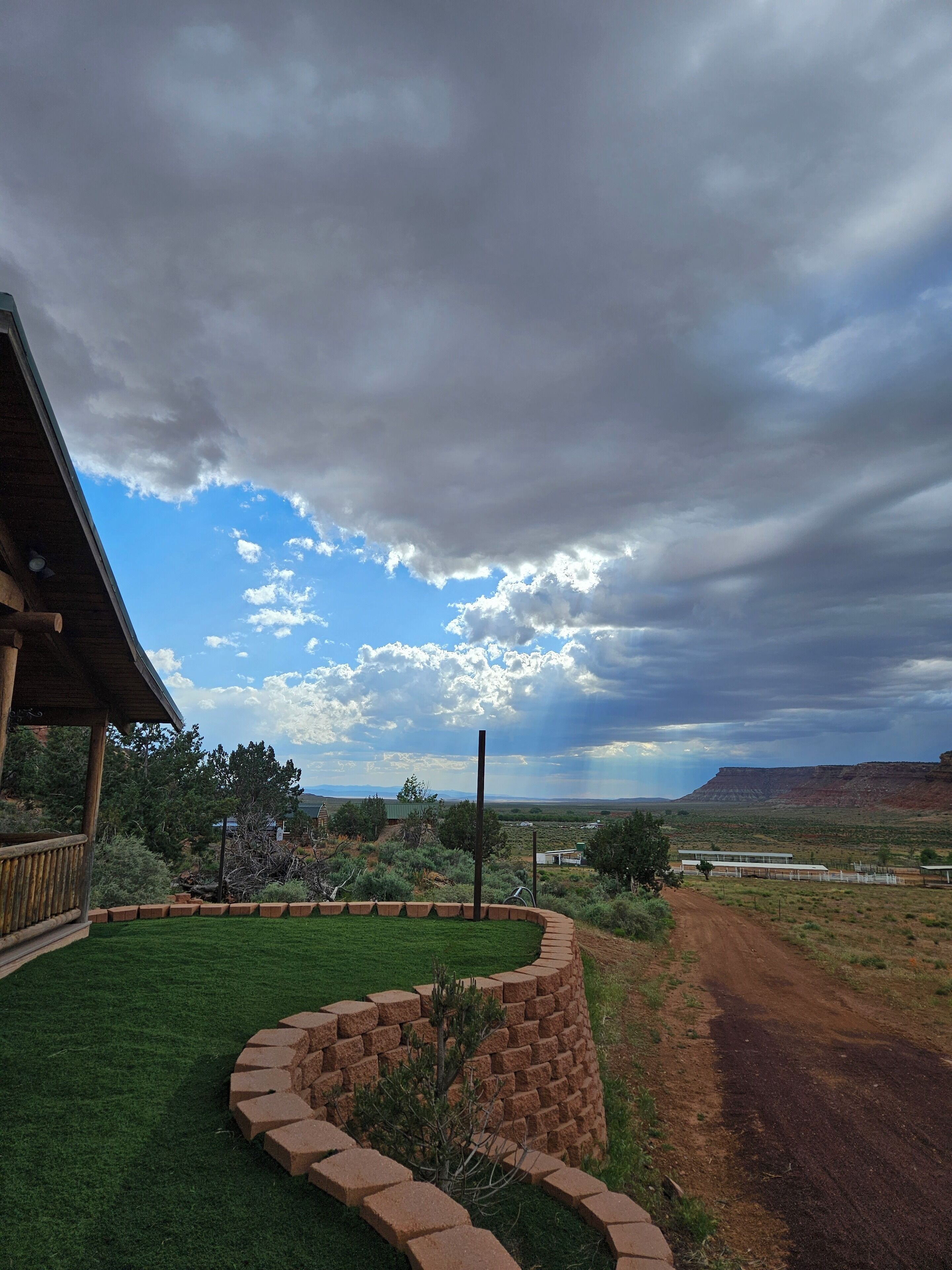 A house with a covered porch, a brick retaining wall, and a dirt path leading to a mountainous landscape.