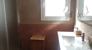 Une salle de bain avec une fenêtre, un miroir, un lavabo et un tabouret en bois.