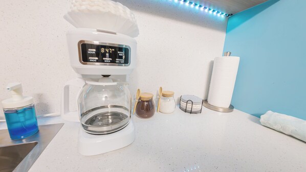 Una cafetera sobre una encimera de cocina, un reloj digital que marca las 8:47 de la mañana, una botella de agua azul y dos frascos con tapa.