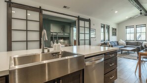 A modern kitchen with a stainless steel sink, a dishwasher, and a large island. There is a living area with a sofa and a dining table.