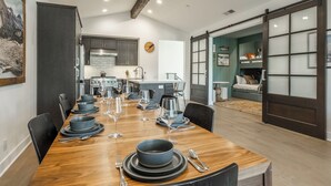 A modern dining room with a wooden table set for six, a kitchen in the background, and a sliding glass door leading to a bedroom.