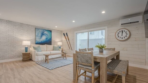 Un salon moderne avec un mur en briques blanches, une grande peinture abstraite, une table à manger en bois avec des chaises en osier et une horloge au mur.