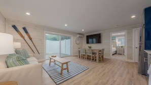 Un vaste espace de vie comprenant un canapé, une table basse et une table à manger avec des chaises. Un téléviseur est fixé au mur, il y a une horloge et une porte-fenêtre coulissante qui donne sur un espace extérieur délimité par une clôture.