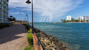 A coastal walkway with a stone railing, a clear sky, and buildings in the background. - Mesmerizing Ocean View King Bed Near Beaches (San Juan)