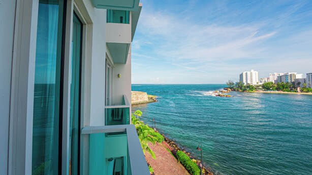A coastal hotel with a balcony overlooking the sea, a paved walkway, and a view of the beach and cityscape. - Mesmerizing Ocean View King Bed Near Beaches (San Juan)