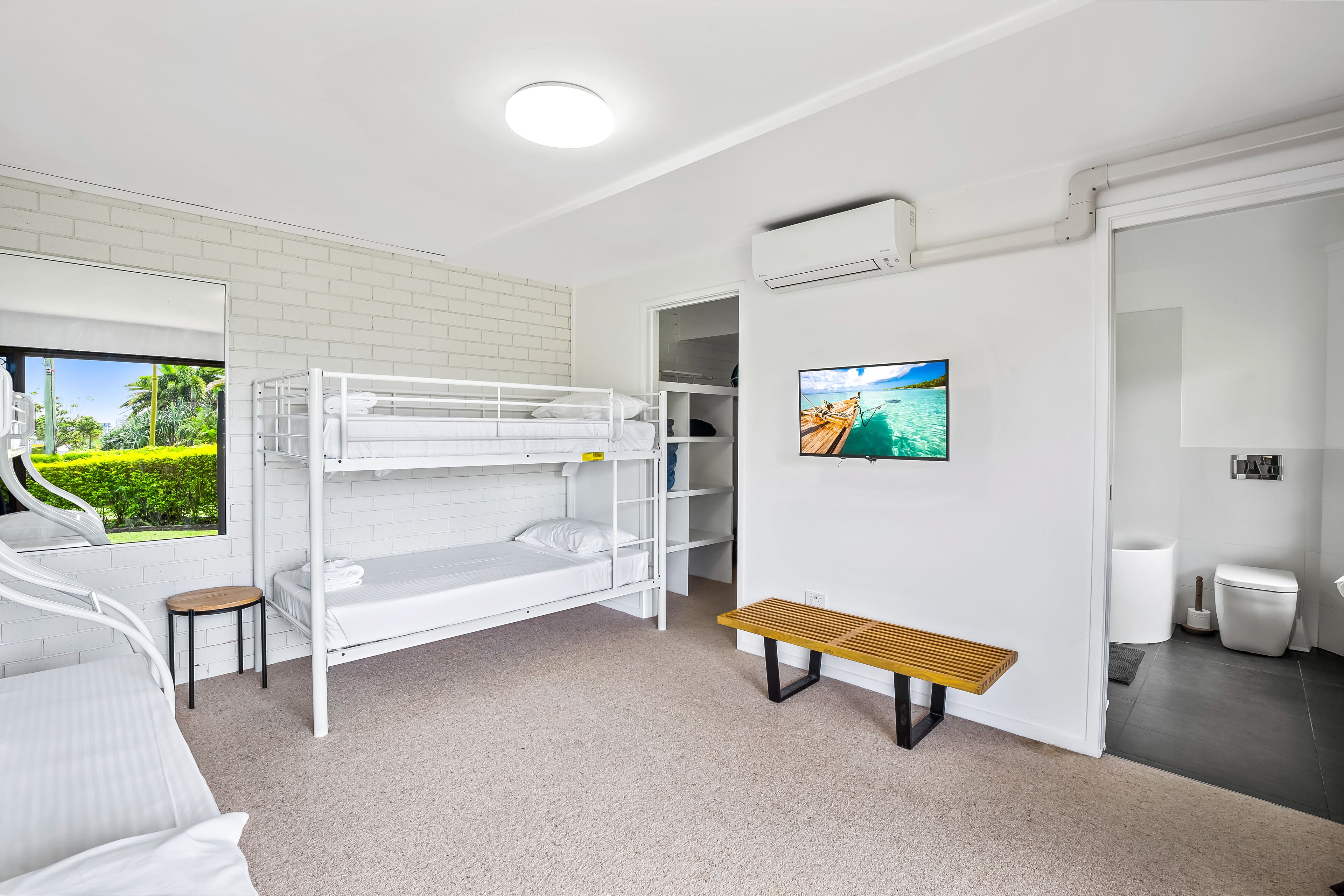 A modern, well-lit room with a bunk bed, a small table, a bench, a TV, and a window with a view of greenery.