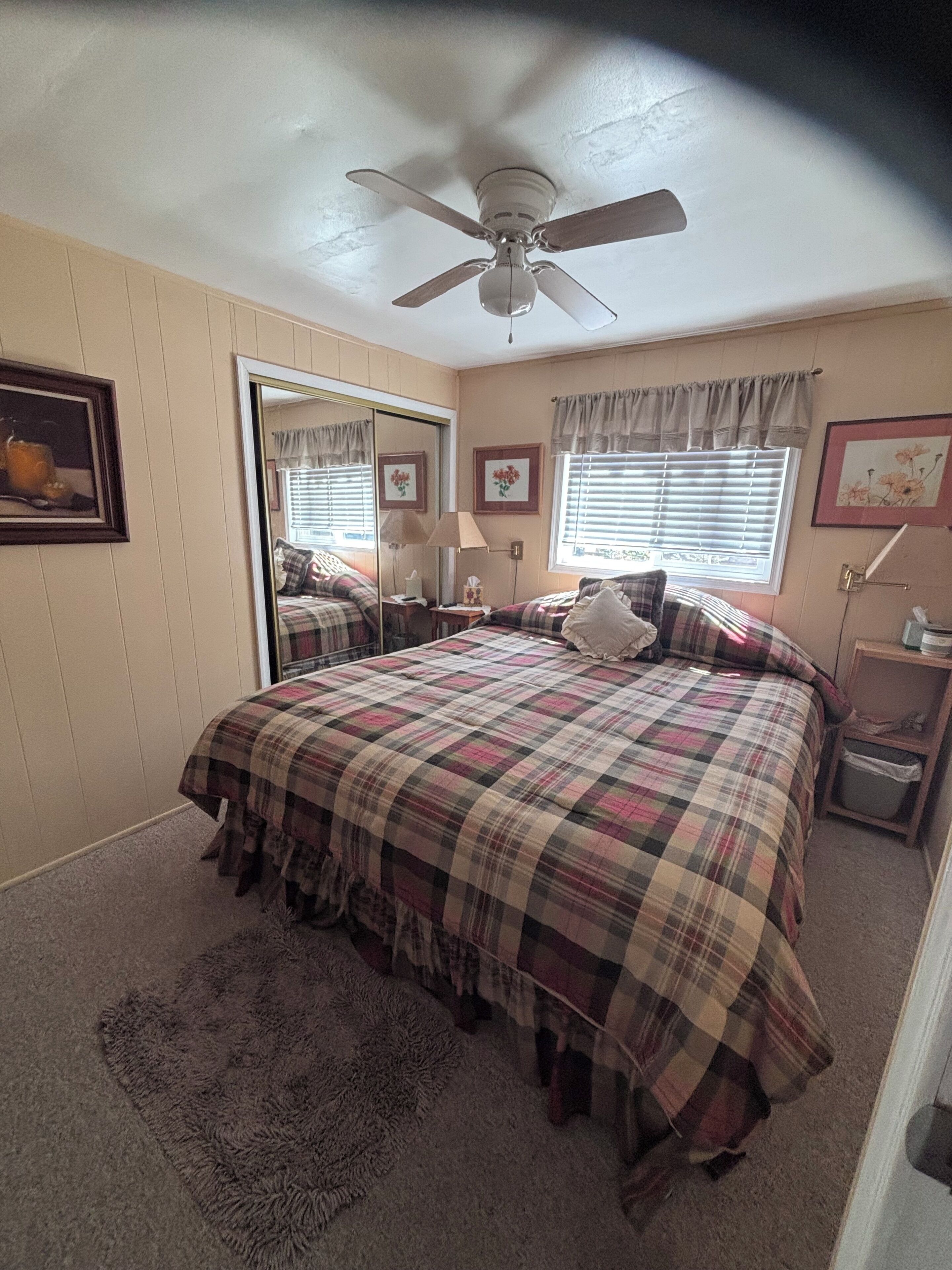 Une chambre à coucher avec un lit, un ventilateur de plafond, une fenêtre avec des stores, un miroir et une table de chevet.