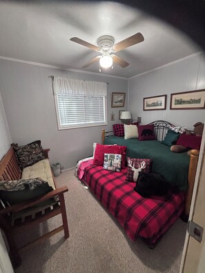 A bedroom with a bed, a chair, a ceiling fan, a window with blinds, and wall-mounted pictures.