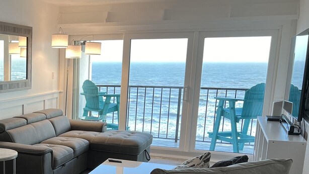 A living room with a leather sofa, a small table, and a TV. There are two chairs and a table outside with a view of the ocean. - Beachfront condo with stunning ocean views. Pet-Friendly! (Galveston)