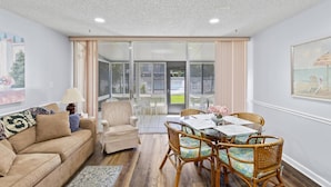 Una sala de estar con un sofá color beige, un sillón pequeño, una mesa de comedor de vidrio con sillas de mimbre, y vistas a un área exterior con patio y piscina.
