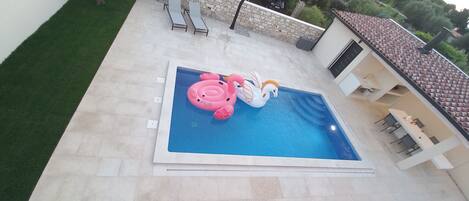 A swimming pool with inflatable toys, a patio area with chairs, and a building with a tiled roof.