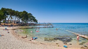 Une plage à l’eau claire, où l’on peut voir des gens nager et prendre des bains de soleil, une jetée en bois et des arbres en arrière-plan.