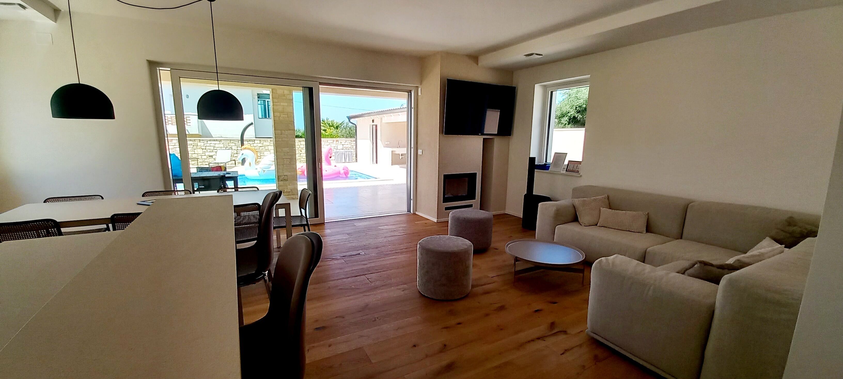 A modern living room with a sofa, ottomans, a dining table with chairs, and a TV mounted on the wall.