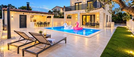 A swimming pool with lounge chairs and inflatable toys, surrounded by a well-maintained lawn and a two-story building with outdoor lighting.