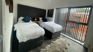 A bedroom with two beds, a nightstand, a lamp, and a view of the outside through a window with blinds.
