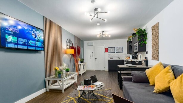 Una sala moderna con un sofá gris, cojines amarillos, una mesa de centro con revistas, un televisor en la pared y una zona de cocina al fondo.