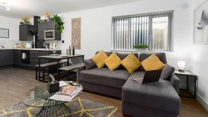 Una sala moderna con un sofá gris, una mesa de centro de vidrio y un comedor con una cocina al fondo.