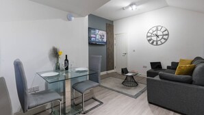 A modern living room with a glass dining table, grey sofa, and a wall-mounted clock. - NH - F3 · Nottage House Premier Apartments (Cardiff)
