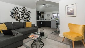 A modern living room with a grey sofa, a yellow armchair, and a coffee table. A kitchen is visible in the background. - NH - F3 · Nottage House Premier Apartments (Cardiff)