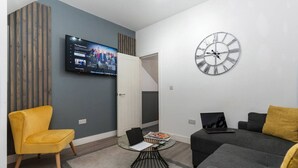 A modern living room with a flat-screen TV, a round wall clock, and a sofa. - NH - F3 · Nottage House Premier Apartments (Cardiff)