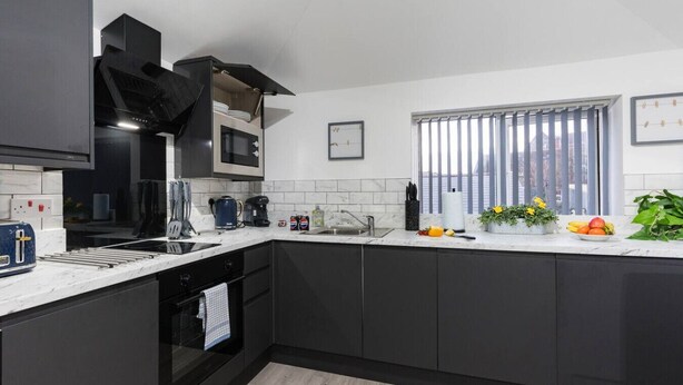 A modern kitchen with dark cabinets, a white countertop, and a window with blinds. - NH - F3 · Nottage House Premier Apartments (Cardiff)