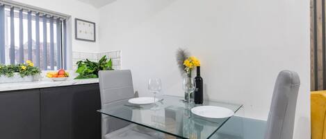 A modern kitchen with a glass dining table, grey chairs, and a small plant arrangement on the counter.