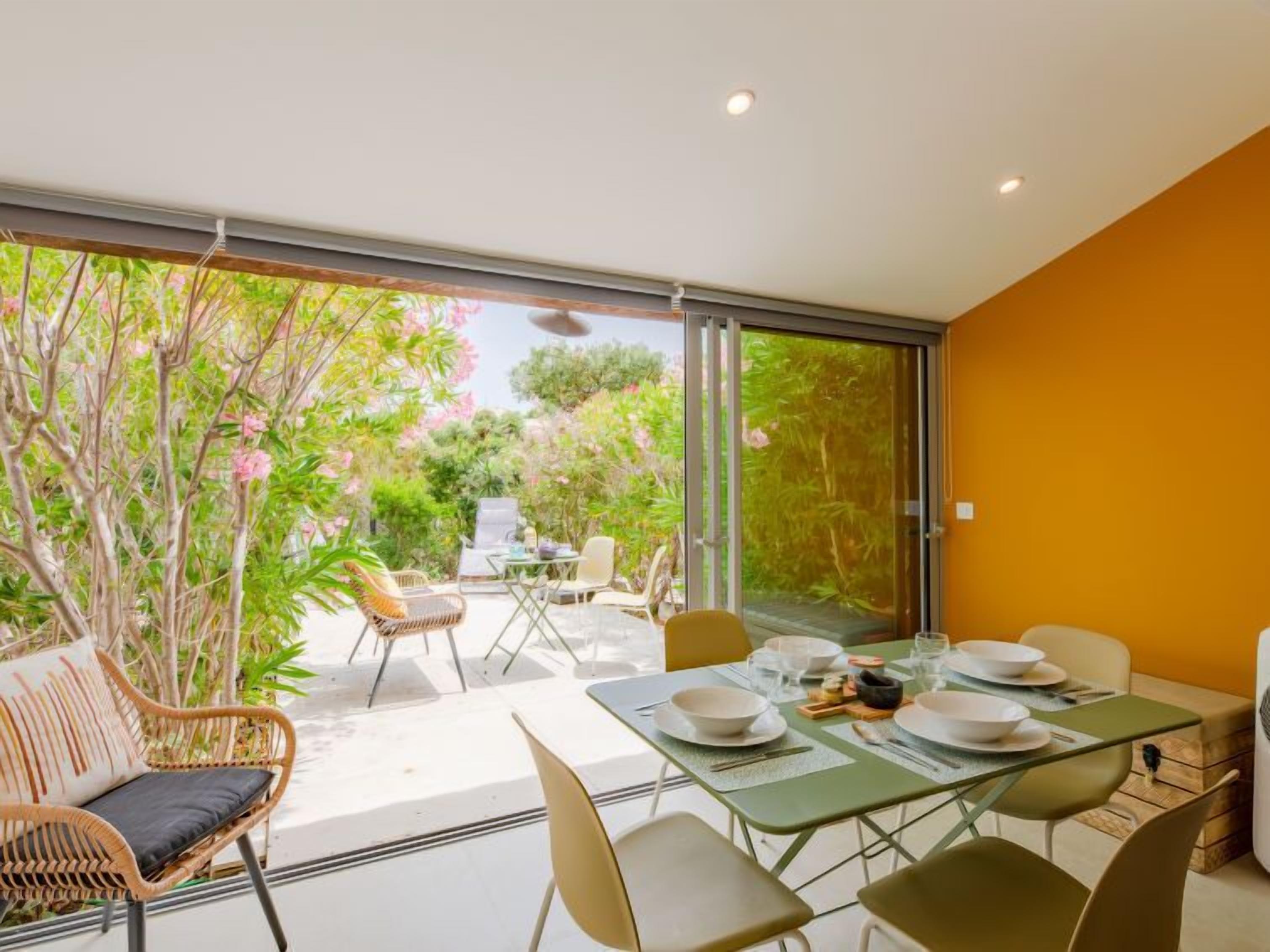 A dining area with a table set for four, surrounded by chairs, and a view of a garden through large glass doors.