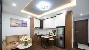 Een compacte keuken met een koelkast, een eethoek met krukken, een kleine tafel met een bloemenvaas, en een bank met een tijdschrift.