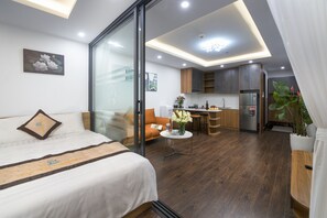 A modern hotel room with a bed, a sofa, a dining table, and a kitchen area.