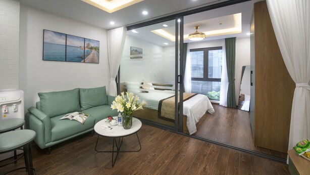 Een moderne hotelkamer met een groene bank, een klein rond tafeltje met een bloemenvaas, een bed met een witte dekbedovertrek en een grote spiegel die de kamer weerkaatst.