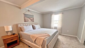 Un dormitorio con una cama grande, una mesita de noche, un cuadro en la pared y una ventana con cortinas.