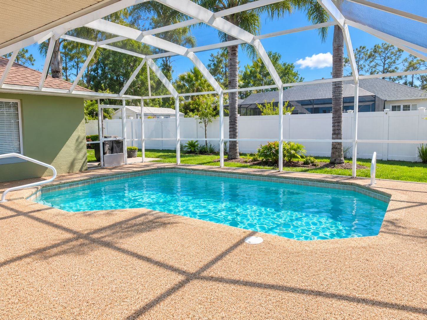 A swimming pool with a covered area, surrounded by a white fence, palm trees, and a clear sky.