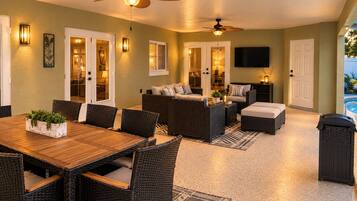 A spacious indoor area with a dining table, wicker furniture, ceiling fans, and a TV mounted on the wall.